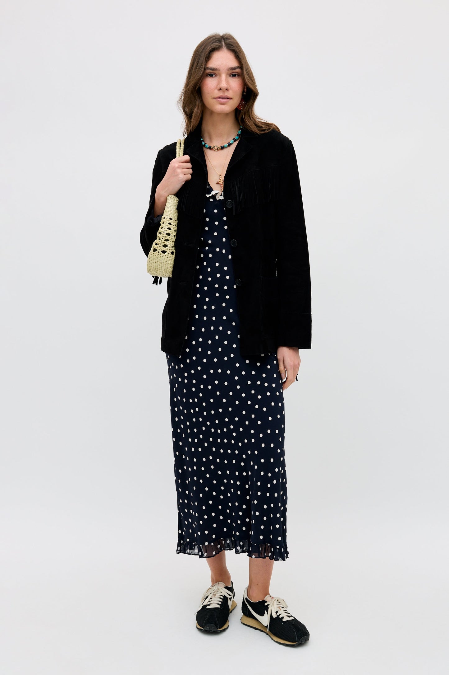 A woman faces forward, wearing the Bethea - Layla Dot Navy dress, paired with a black jacket, black sneakers, a small woven bag, loose wavy hair, and a beaded necklace.