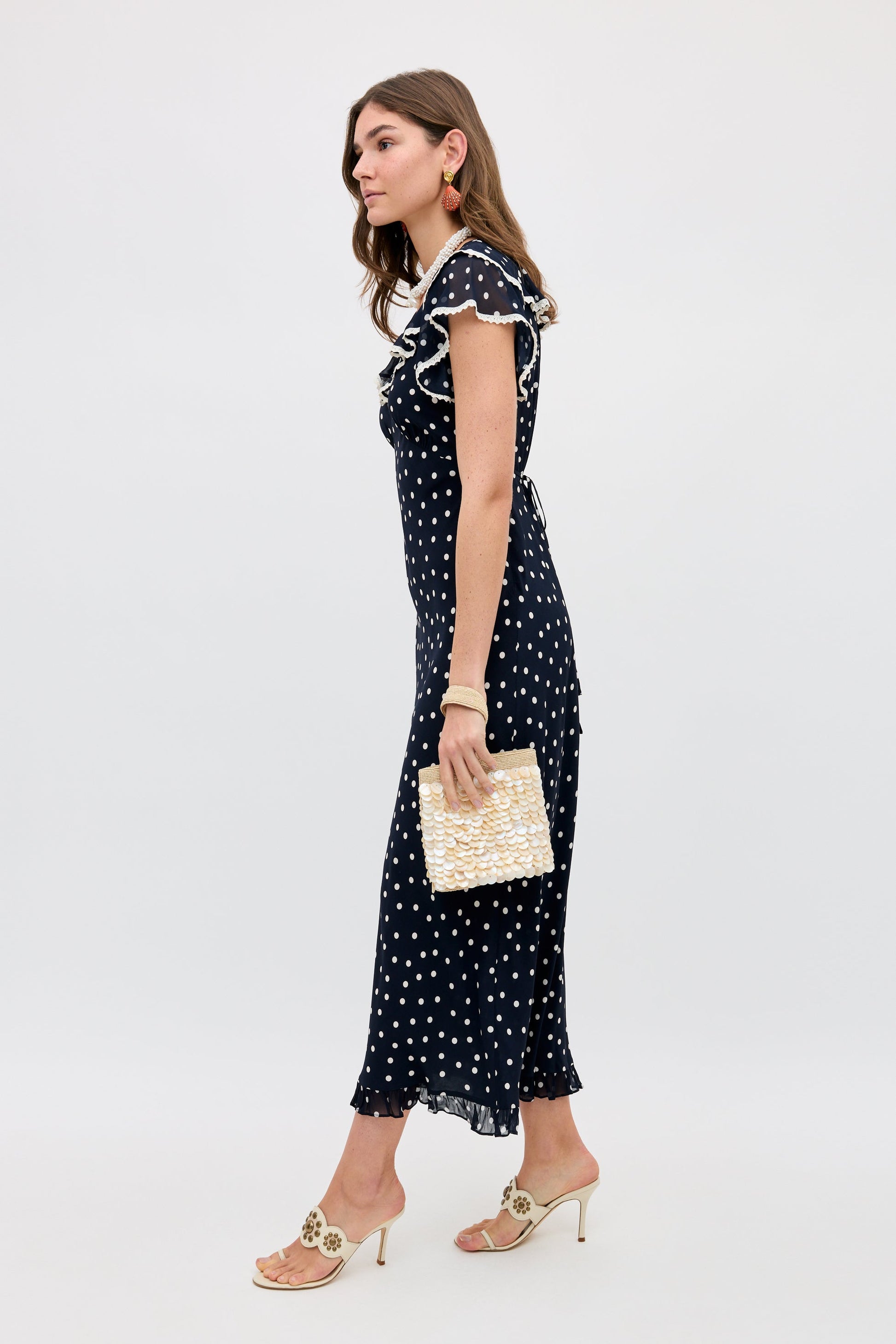 A woman wearing the Bethea - Layla Dot Navy midi dress with short sleeves and ruffle details, paired with a woven clutch and heeled sandals. She has long hair and is posed against a simple light background.