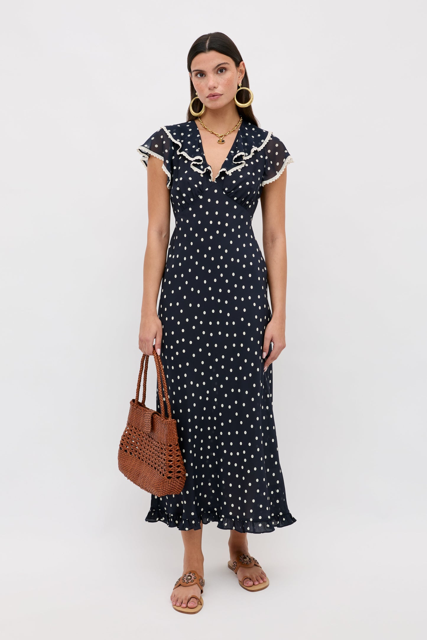 A woman wears the Bethea - Layla Dot Navy dress, featuring short sleeves, polka dots, and a ruffled collar. She holds a brown woven handbag, wears large hoop earrings and flat sandals, standing against a plain white background.